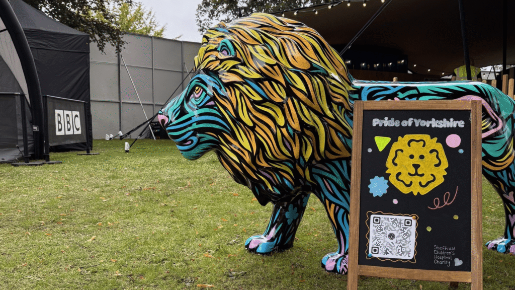 An image taken at Tramlines Festival 2025. A painted lion faces the left of the image, next to him is a wooden chalk board which reads 'Pride of Yorkshire' and in the background you can see the BBC Radio Sheffield tent.