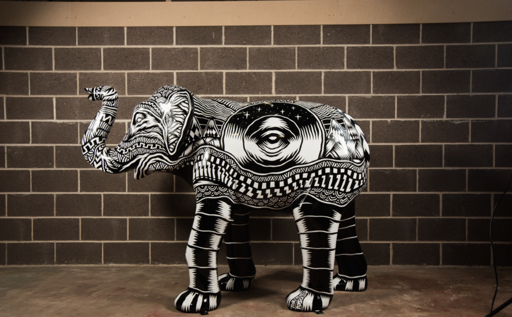 An image of an Elephant sculpture against a brick wall. The elephant features an all-over black and white, psychedelic design and draws inspiration from vinyl records.