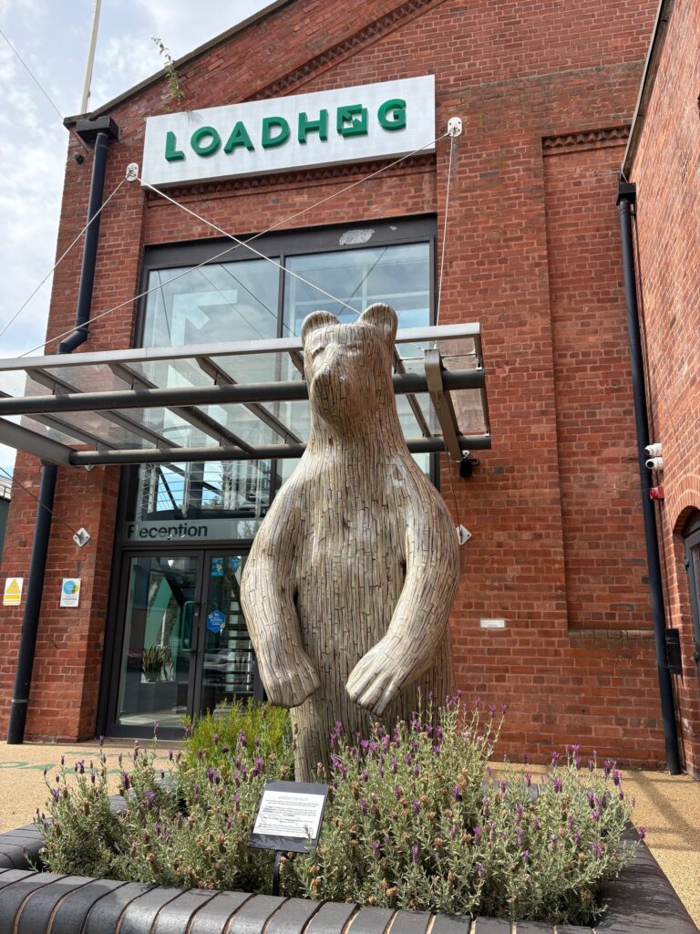 An image of a bear sculpture. It stands proudly in a planet surrounded by lavender, outside of a brick building. The bear's design is made to look like wood, and in the background a sign reads LOADHOG.