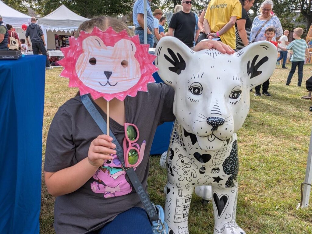Someone stands next to a small lion cub sculpture which features all-over black and white doodles. Their face is covered by a bright pink lion mask.