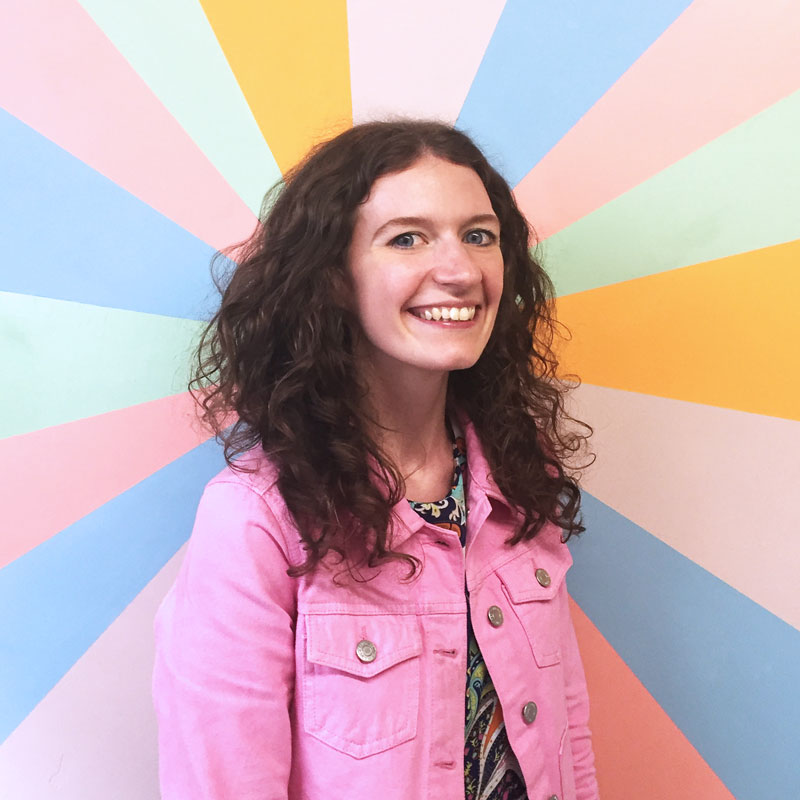 A person in a pink denim jacket with curly dark brown hair stands in front of a rainbow background, smiling at the camera.