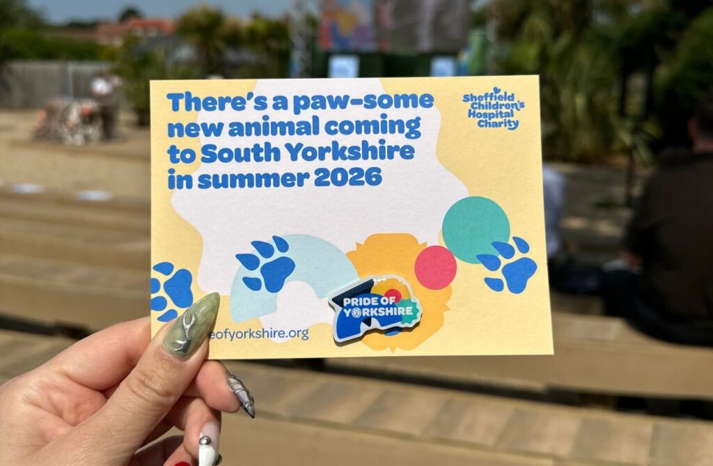 A hand holding a brightly coloured postcard which reads 'there's a paw-some new animal coming to South Yorkshire in summer 2026'