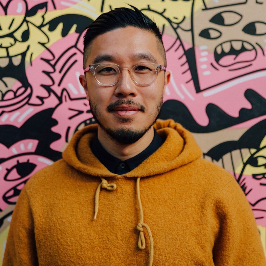 A person with short hair, wearing glasses and an orange hoodie faces the camera. They are stood in front of a vibrant, mural-filled wall.