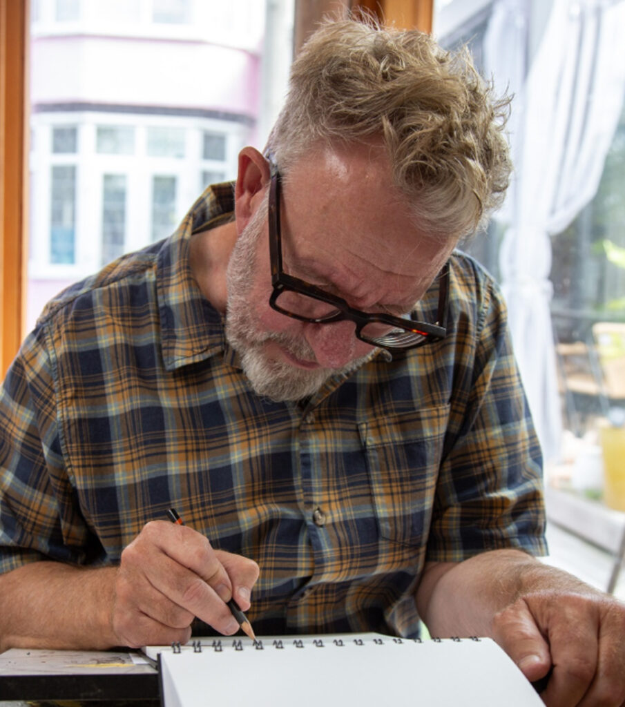 A person wearing glasses and a checked shirt works on a drawing. They are focussed and using a pencil to draw.