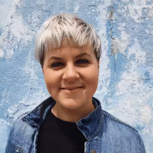 A person with short, bright blonde hair stands against a sky-blue background and smiles at the camera.