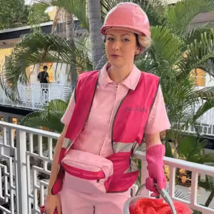 A person wearing all pink - including a hart hat, belt bag and high-vis vest stands central and looks at the camera. They look to be stood on a balcony with a tree in the background.