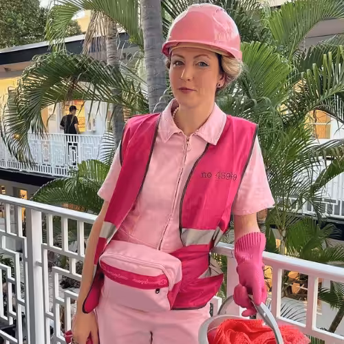 A person wearing all pink - including a hart hat, belt bag and high-vis vest stands central and looks at the camera. They look to be stood on a balcony with a tree in the background.