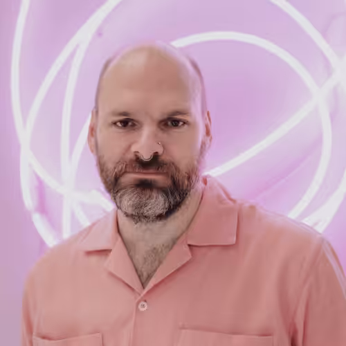 A person with a beard and wearing a pink, button-down shirt looks into the camera. They are stood against a pink, neon background.