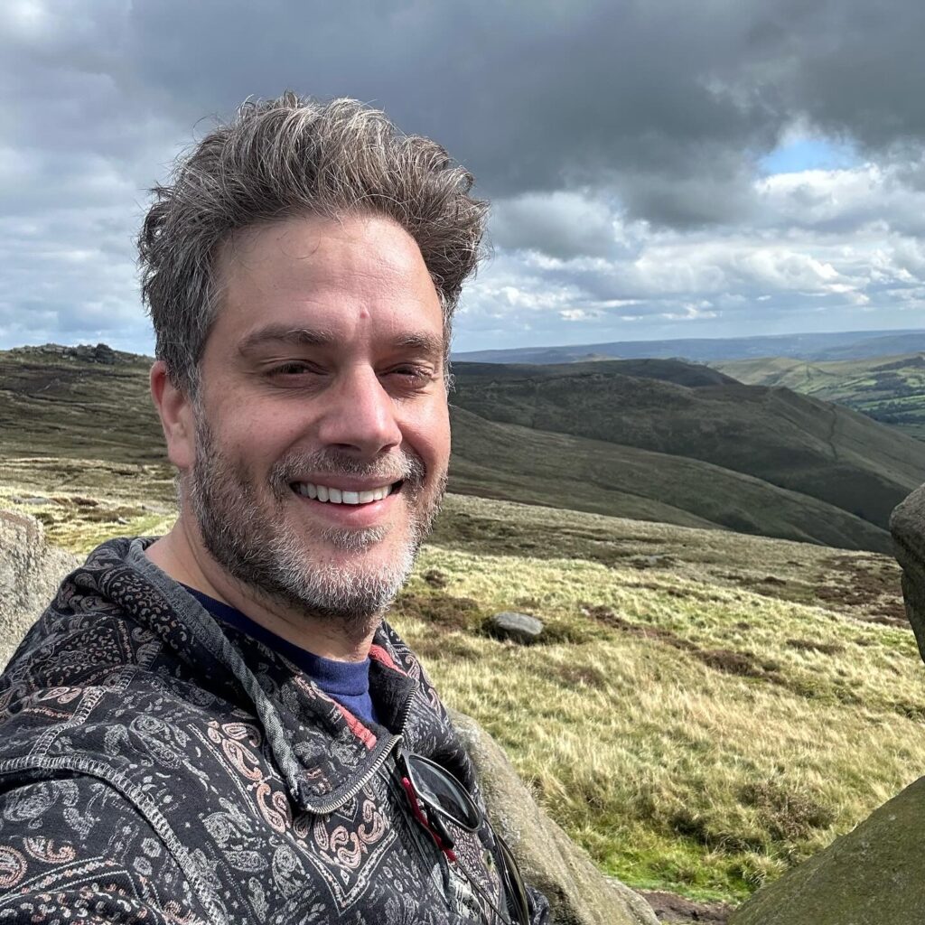 A person takes a picture of themselves in the peak district. It is a vast background with hills and rolling skies. The person is smiling.