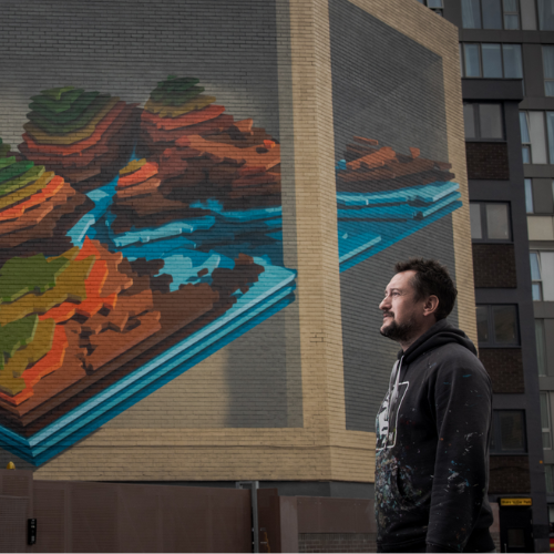 A person stands at a distance from the camera. They have short brown hair and a beard, and are facing the left. Behind them, a mighty, colourful optical illusion mural sits on the side of a brutalist building. Photo by Chris Saunders.