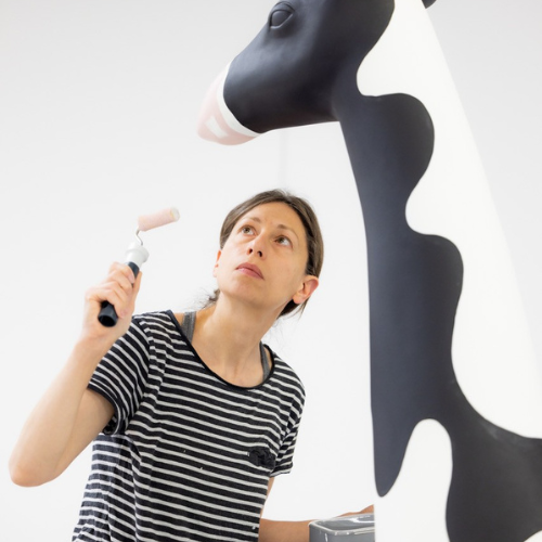 A person with tied-back brown hair wearing a striped shirt stands beneath a sculpture. They are looking up at it and holding a paint roller.