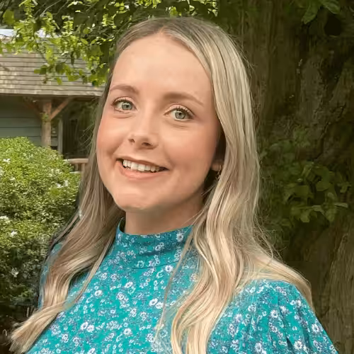 A person with long, blonde hair looks at the camera smiling. They are stood against a tree-filled background and wearing a green dress.,