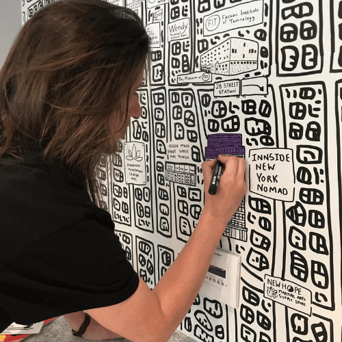 A person with shoulder-length brown hair faces a mural and is adding to its monochrome design.