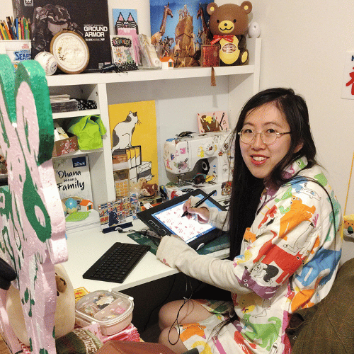 A person sits at a desk and turns to look at the camera. They have long dark hair and wear glasses. Their desk is filled with artwork, and they are using a sketch pad.