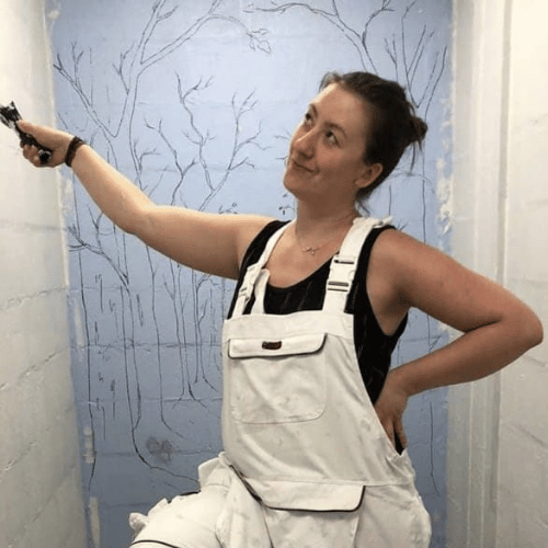 A person in white dungarees stands central in the image. They have one hand on their hip, and the other wields a paintbrush. They look to be stood in front of and next to a mural which is yet to be painted.