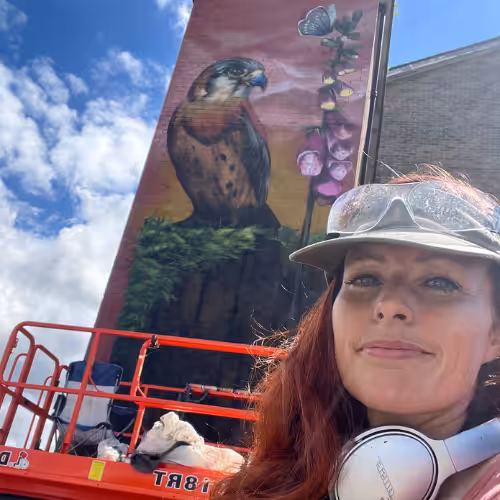 A person takes a selfie in front of a multi storey mural of a bird. The sun is shining and the sky is blue, and they wear a baseball cap and headphones.