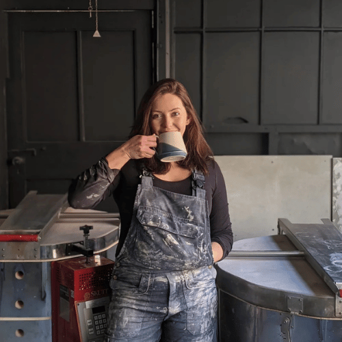 A person with shoulder-length brown hair and wearing paint-covered dungarees stands in a room which looks like a workshop. They are facing the camera and holding a mug of tea to their mouth, with the other hand in a pocket.