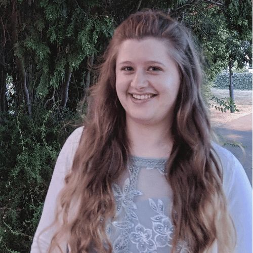 A person with long brown hair smiles at the camera. They are wearing a light blue dress and are stood in front of a tree.