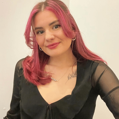 A person with pink, shoulder-length hair and wearing a black dress holds the camera with one hand and smiles towards it.
