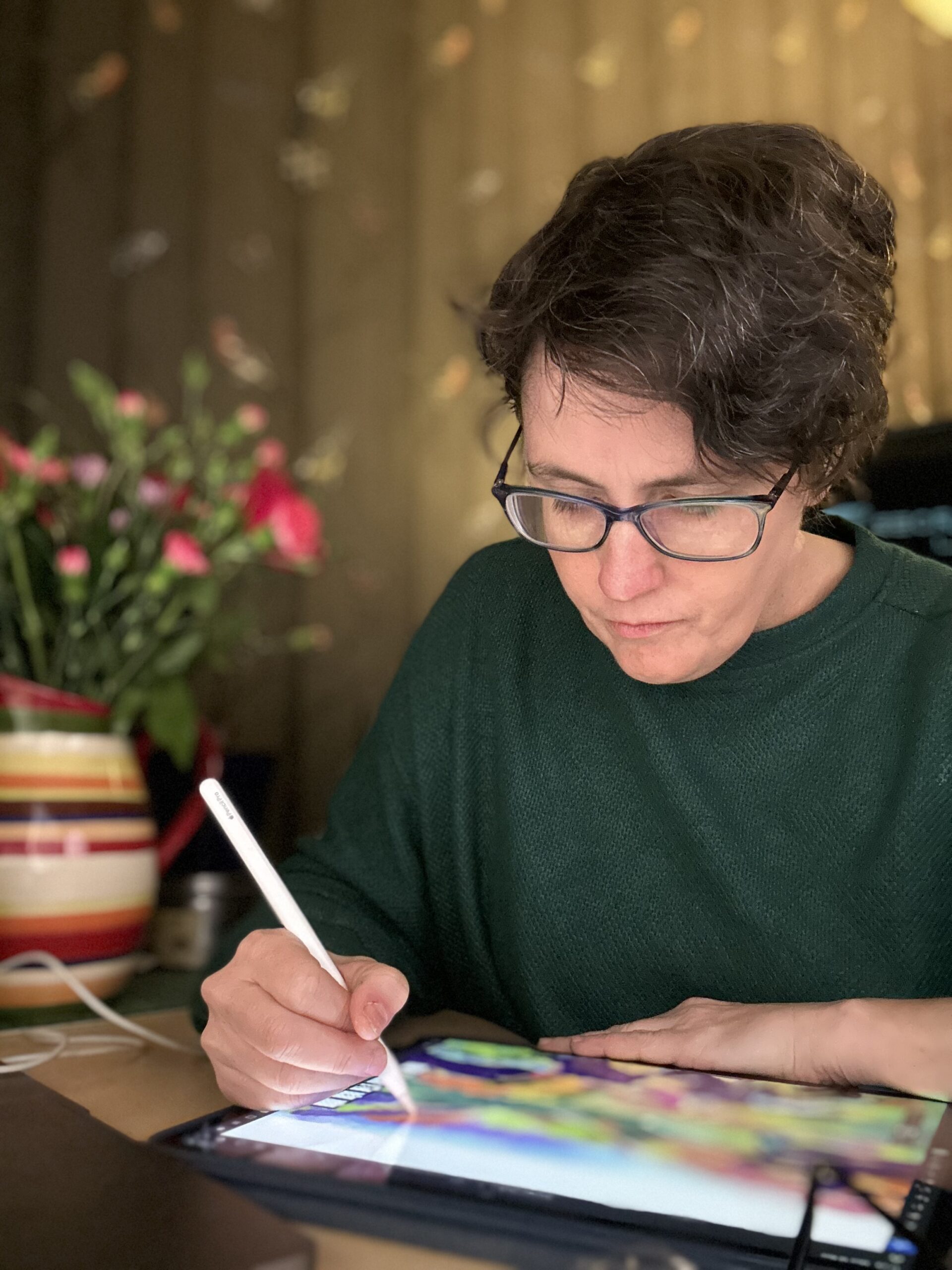 A person with short brown hair and a dark green jumper paints a landscape on an ipad in a dark room with flowers in a stripy vase.