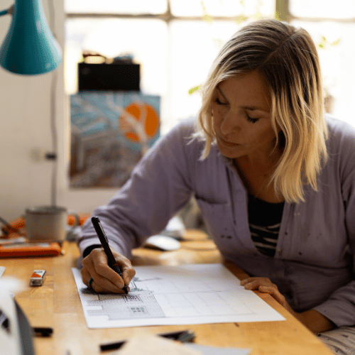 A person with shoulder-length blonde hair works at a desk on a piece of artwork. The sun comes into the room from the window behind.