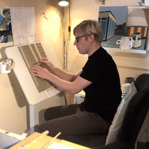A person with short blonde hair, wearing glasses and all-black works at a desk beneath a lamp on a piece of art.