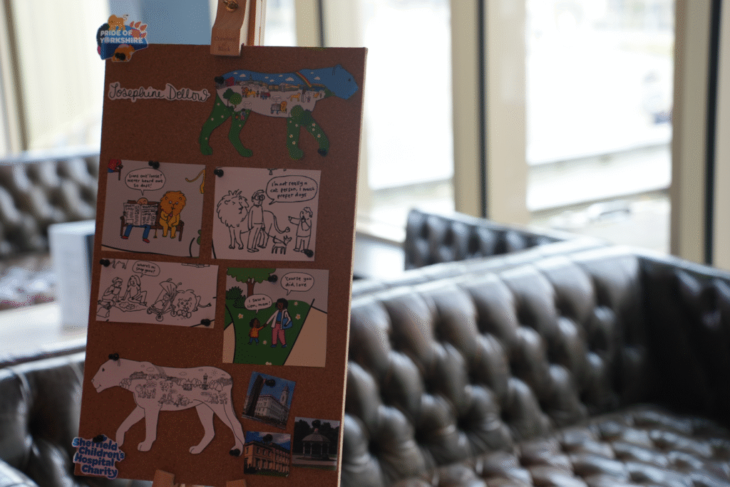 A photograph of a cork board which features a Pride of Yorkshire lioness design. There are two cut-outs of the lioness template, with one side appearing in full colour and a Barnsley skyline design, and the other is black and white with a second Barnsley skyline themed design. The artist is Josephine Dellow and her name can be seen in the top corner.