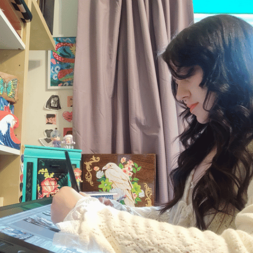 A person with long dark hair and wearing a cream jumper works at a desk. They are side on to the camera and are illustrating something.
