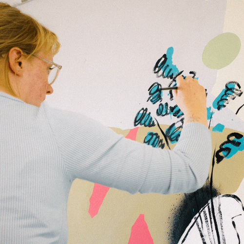 A person with blonde hair and wearing glasses is facing away from the camera so only the side of their face is seen. They are working on a piece of art - painting blue florals.
