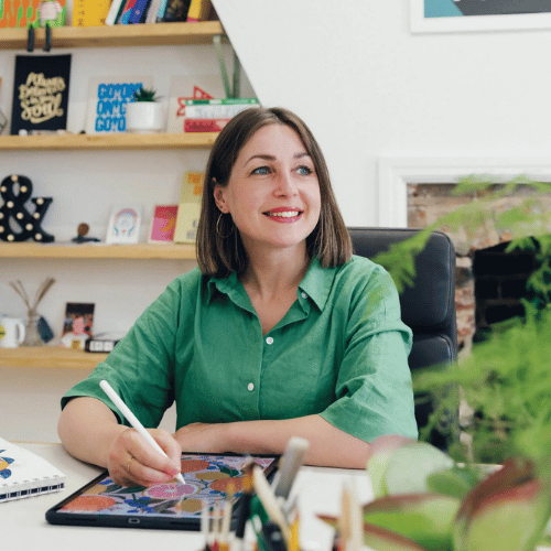 A person with shoulder-length brown hair and wearing a green button-up shirt sits at a desk and looks off camera. They are working on a piece of art at a desk.