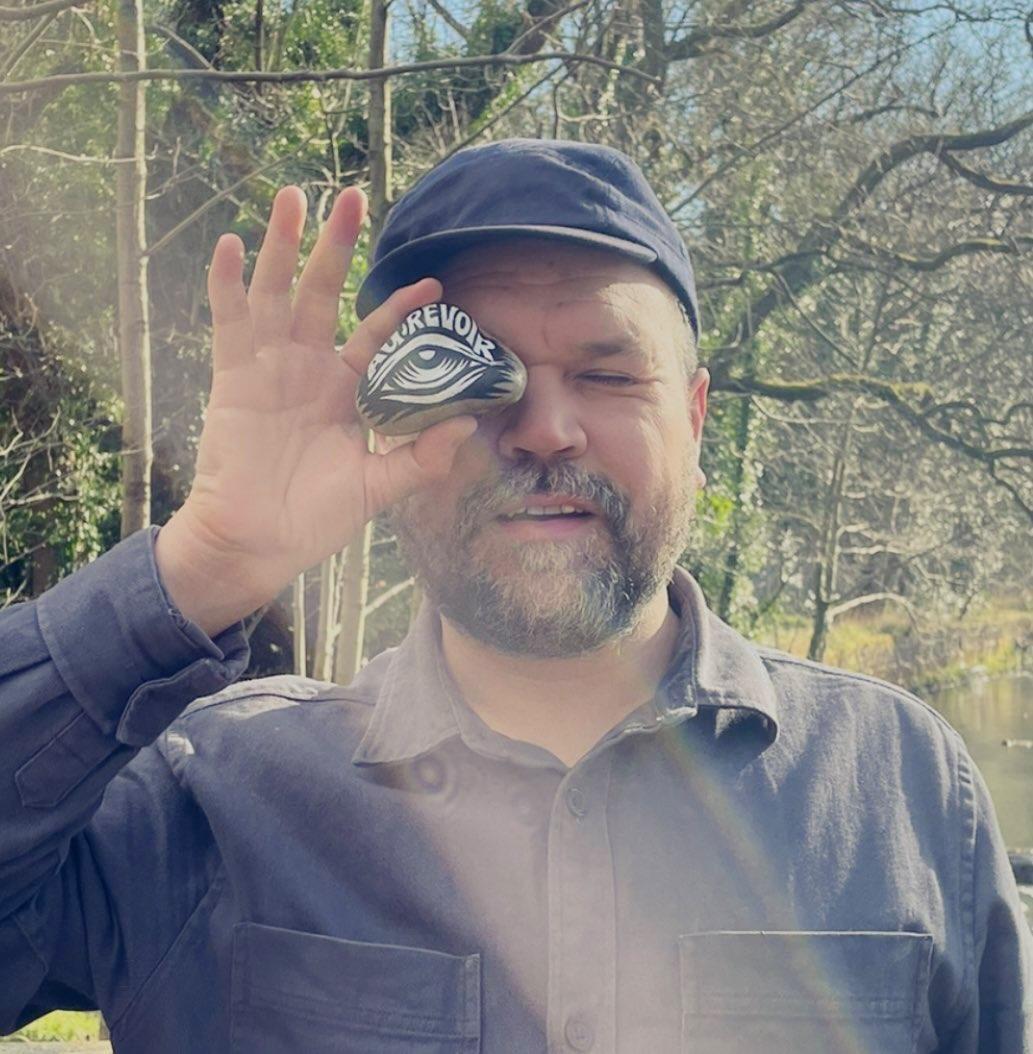 A picture of a person with a beard and wearing a hat facing the camera. They hold an illustrated circle over their eye and the sun shines.