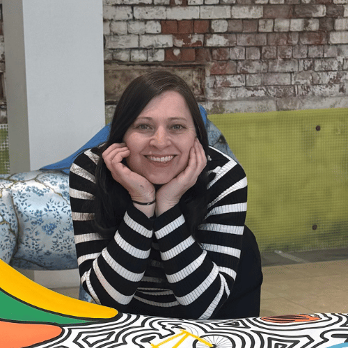 A person with brown hair and wearing a monochrome stripey shirt holds their chin in their hands and smiles at the camera. In the foreground, a hint of a sculpture can be seen.