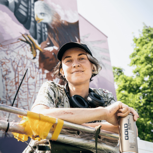 A person with short hair tucked into a baseball cap looks down at the camera. They hold a can of spray paint in their hand, and behind them you can see a soaring mural.