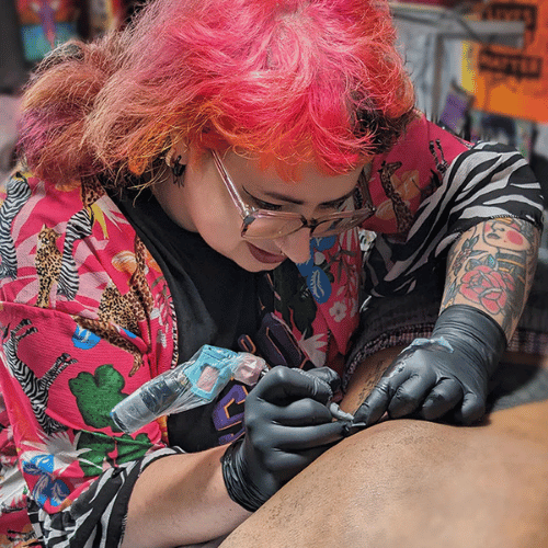 A person with bright pink hair and glasses is focussed on tattooing. They look down at their subject.