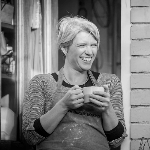 A black and white picture of a person with short hair who wears an apron over a jumper. They are looking off to the right of the camera, smiling widely and holding a mug of tea.