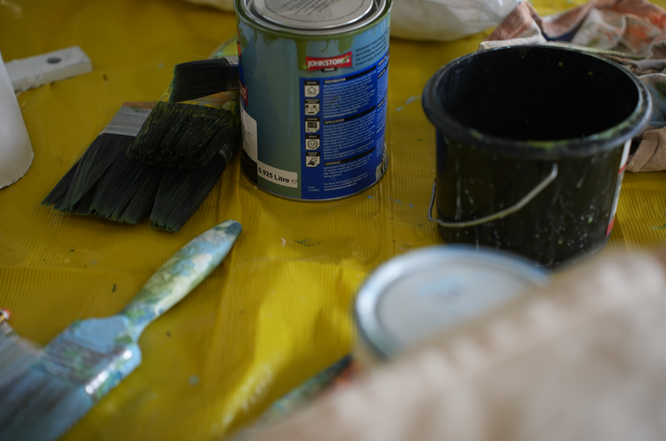 An image which shows a yellow tarp-like flooring with paint pots, brushes and buckets on top of it. The brushes are covered in paint, like they've been put down for only a moment!