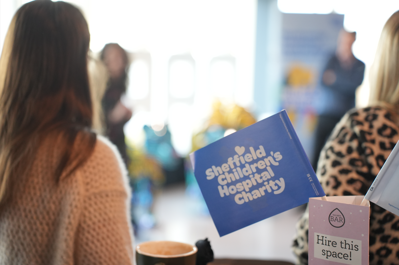 A photograph taken at an event. In the distance you can see brightly coloured sculptures and someone delivering a speech. In the foreground, the back of someone's head as they listen, and a blue flag that says Sheffield Children's Hospital Charity.