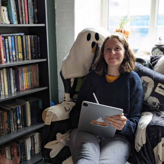 A person sits in an armchair holding a tablet and facing the camera. There's a bookshelf to their left and a ghost cushion behind them.