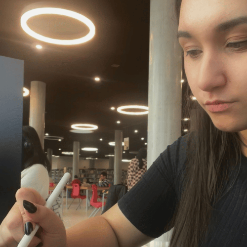 A person with long brown hair is close to the camera, looking down at a piece of art they're working on. 