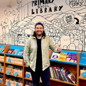 A photograph of a person wearing a grey jacket and a black t-shirt in front of an illustrated wall. They are smiling and giving a thumbs up.