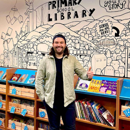 A photograph of a person wearing a grey jacket and a black t-shirt in front of an illustrated wall. They are smiling and giving a thumbs up.