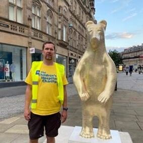 A photo of a person stood wearing a Sheffield Children's Hospital Charity t-shirt and stood next to a tall bear sculpture.
