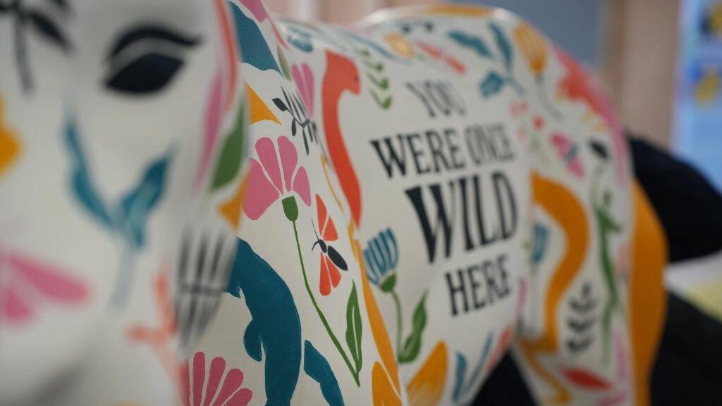 A brightly decorated sculpture can be seen. Its face is close to the camera and out of focus, its body can be seen in the background and is decorated with bright coloured florals. The text reads: You were once wild here.