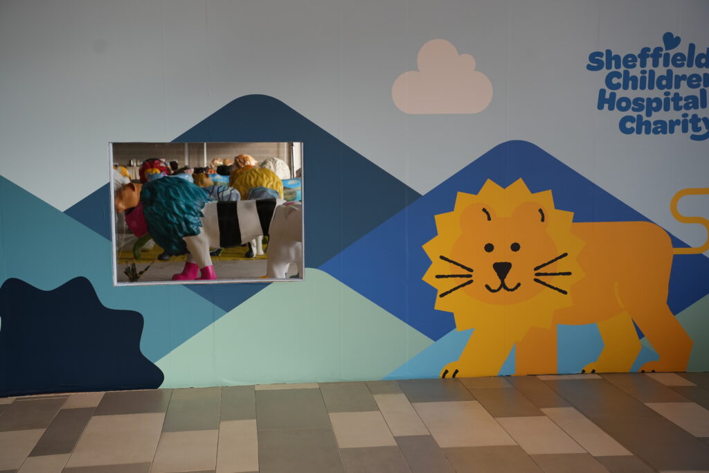 A photograph of the Pride of Yorkshire painting space from the outside. There's a brightly decorated wall, with rolling hills and a cartoon lion, and in the middle of the image there's a window where you can see through to the painting space - it's packed with brightly coloured lions.