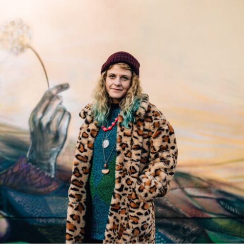 A photo of a person wearing a leopard-print coat and a beanie hat. They have blonde hair with blue at the tips, and are looking at the camera with their hands in their pockets. Behind them, a mural can be seen.