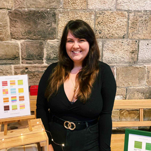 A photograph of a person with long, dark hair and wearing all black. They are smiling at the camera and appear to be stood at a stall where they are selling their artwork.