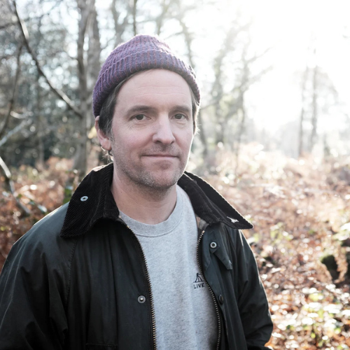 A photograph of a person wearing a beanie hat and a jacket. They are looking at the camera and are stood in a wooded setting - the sun shines through at the back of the photo.