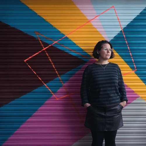 A photograph of a person wearing all-black stood in front of a painted shop shutter. They have their hands in their pockets and are looking over to the left.