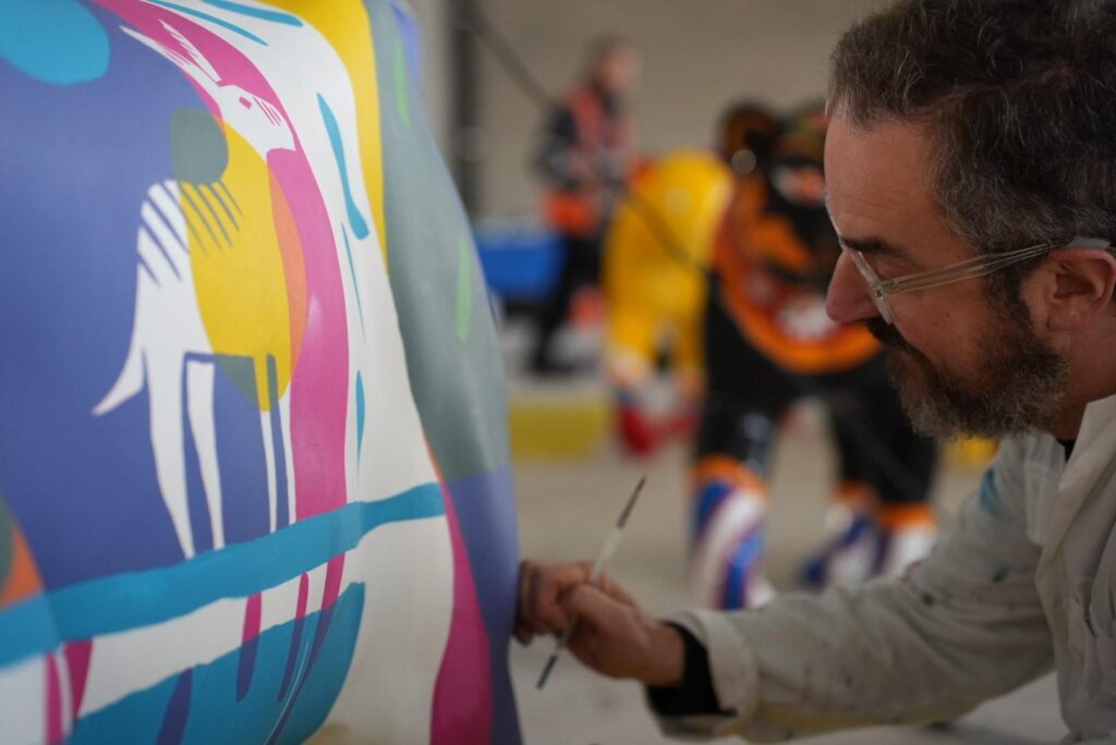 A person in a white boilersuit works on a lion sculpture. They are wearing glasses and hold a paint brush.