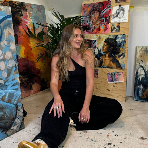 A picture of a person with long, blonde hair wearing all black sat on the floor. They look off to the left and smile at something off camera. Their paintings can be seen behind them.